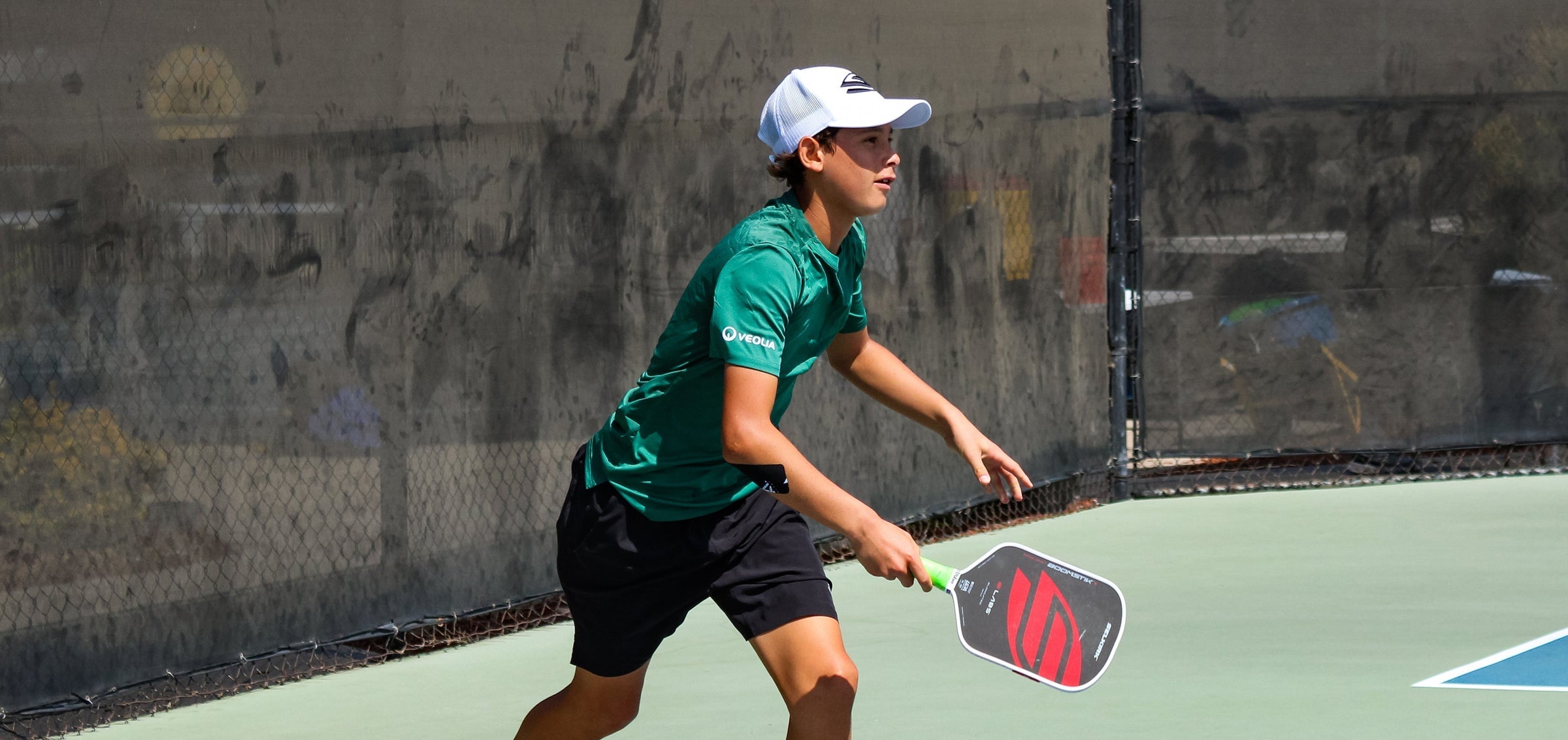 Youth player lunging to return a shot on an outdoor court, wearing green shirt and white cap, holding red-and-black paddle
