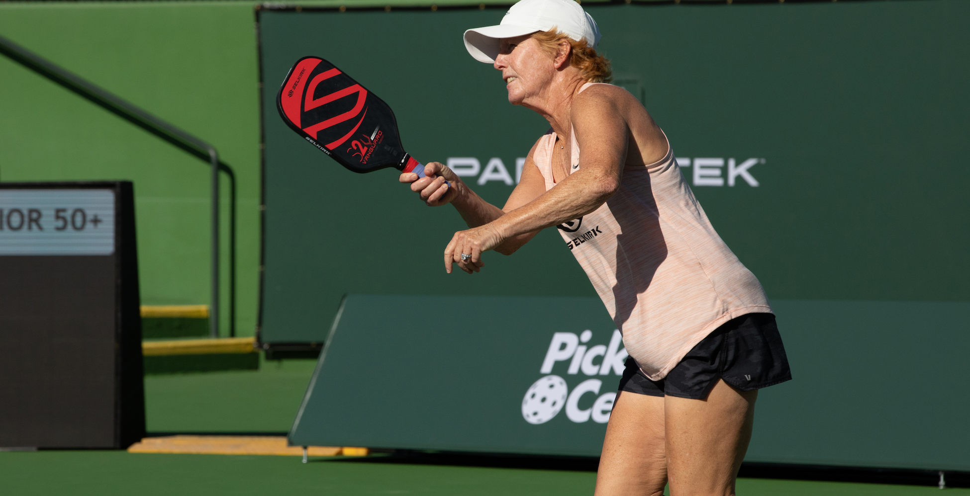 Woman in white cap hitting pickleball with red-black paddle; background signs read Pickle Ce, TEK and scoreboard shows OR 50+