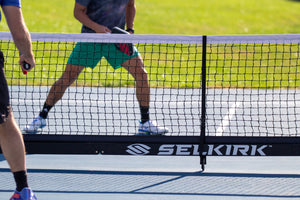 Pickleball players hold paddles on opposite sides of a Selkirk net on an outdoor court