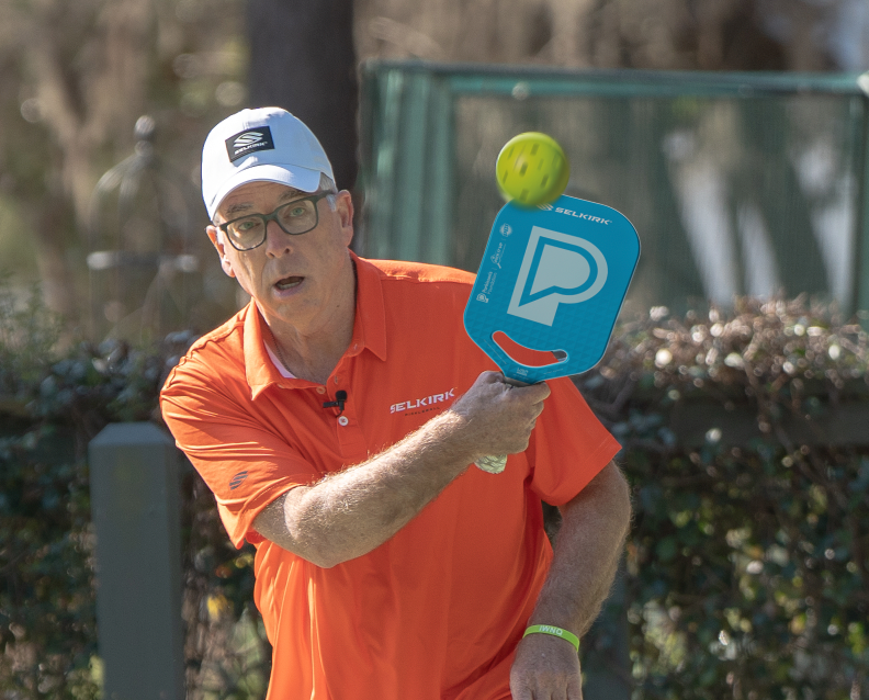 Older man in orange Selkirk polo hitting a yellow pickleball with a blue Selkirk paddle on an outdoor court
