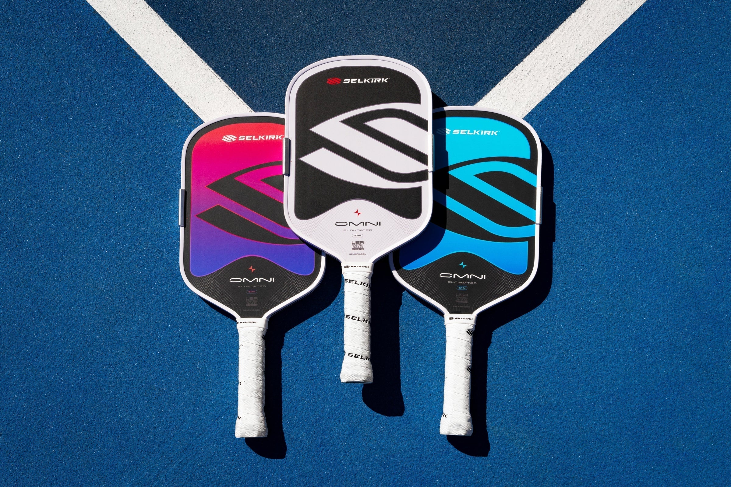 Overhead view of three Selkirk OMNI elongated pickleball paddles in pink, white, and blue on a blue court