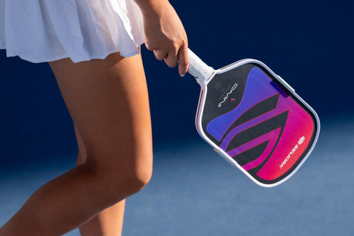 Angled view of Selkirk OMNI pickleball paddle showing purple-pink face graphic and white handle held by player against a blue court background