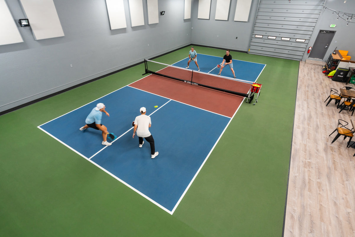 Four players playing doubles pickleball on an indoor court with a Selkirk net and a ball cart near the sideline