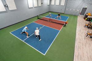 Four players playing doubles pickleball on an indoor court with a Selkirk net and a ball cart near the sideline
