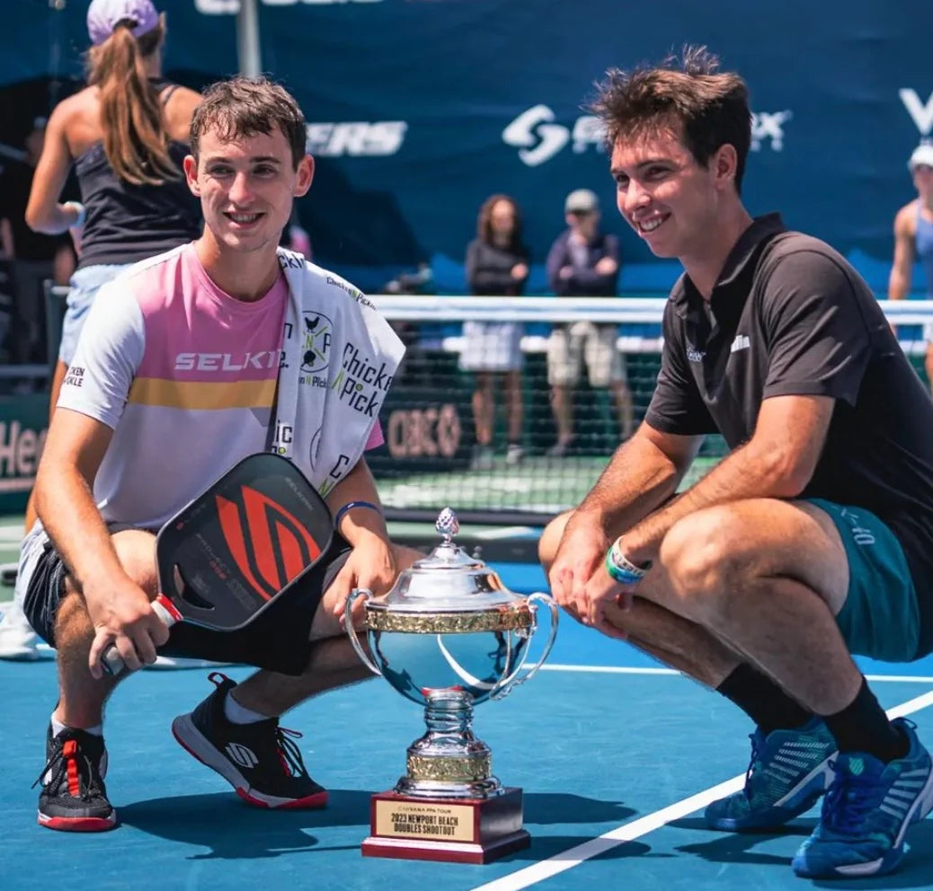 Two pickleball players kneeling beside a trophy on the court after a match.