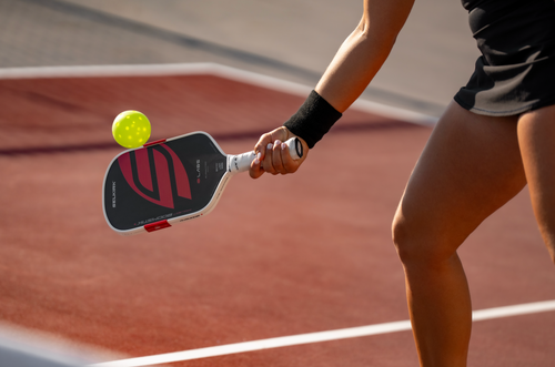 A close up shows a woman hitting a pickleball off a Selkirk Boomstik. 