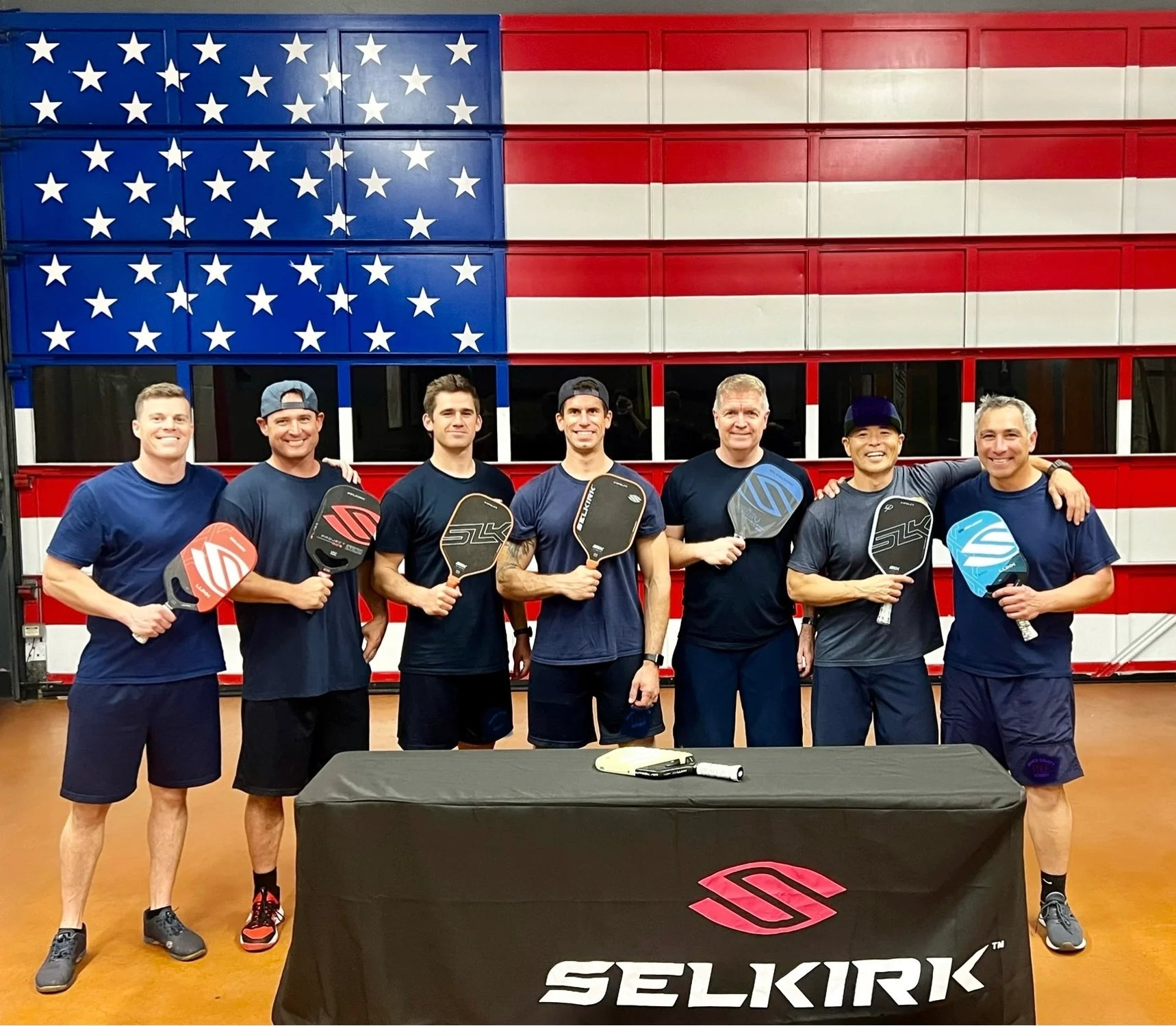 Seven firefighters, including Brian Pingree, pose with Selkirk pickleball paddles in front of an American flag backdrop.