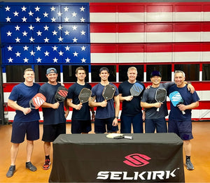 Seven firefighters, including Brian Pingree, pose with Selkirk pickleball paddles in front of an American flag backdrop.