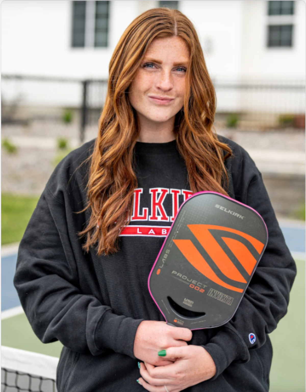 Brooke Bromley holding a pickleball paddle on a court, wearing a black sweatshirt with red lettering.