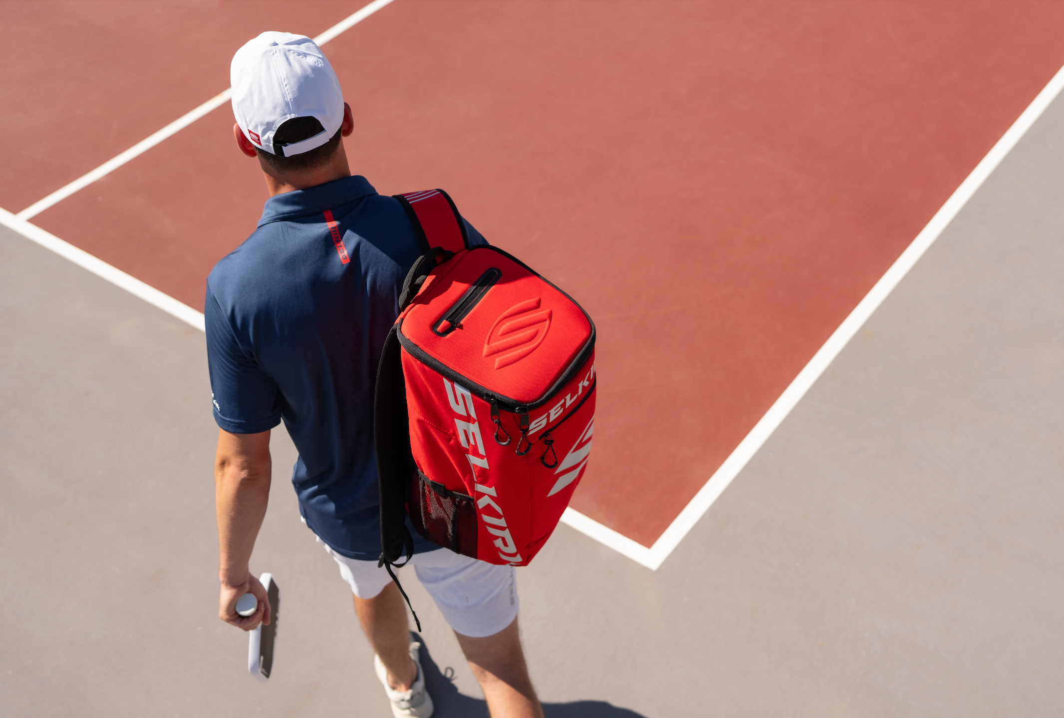 A an image shows a man's back and Selkirk backpack as he crosses around a pickleball court. 