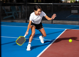 A woman smiles as she prepares to dink a pickleball on an indoor court. 