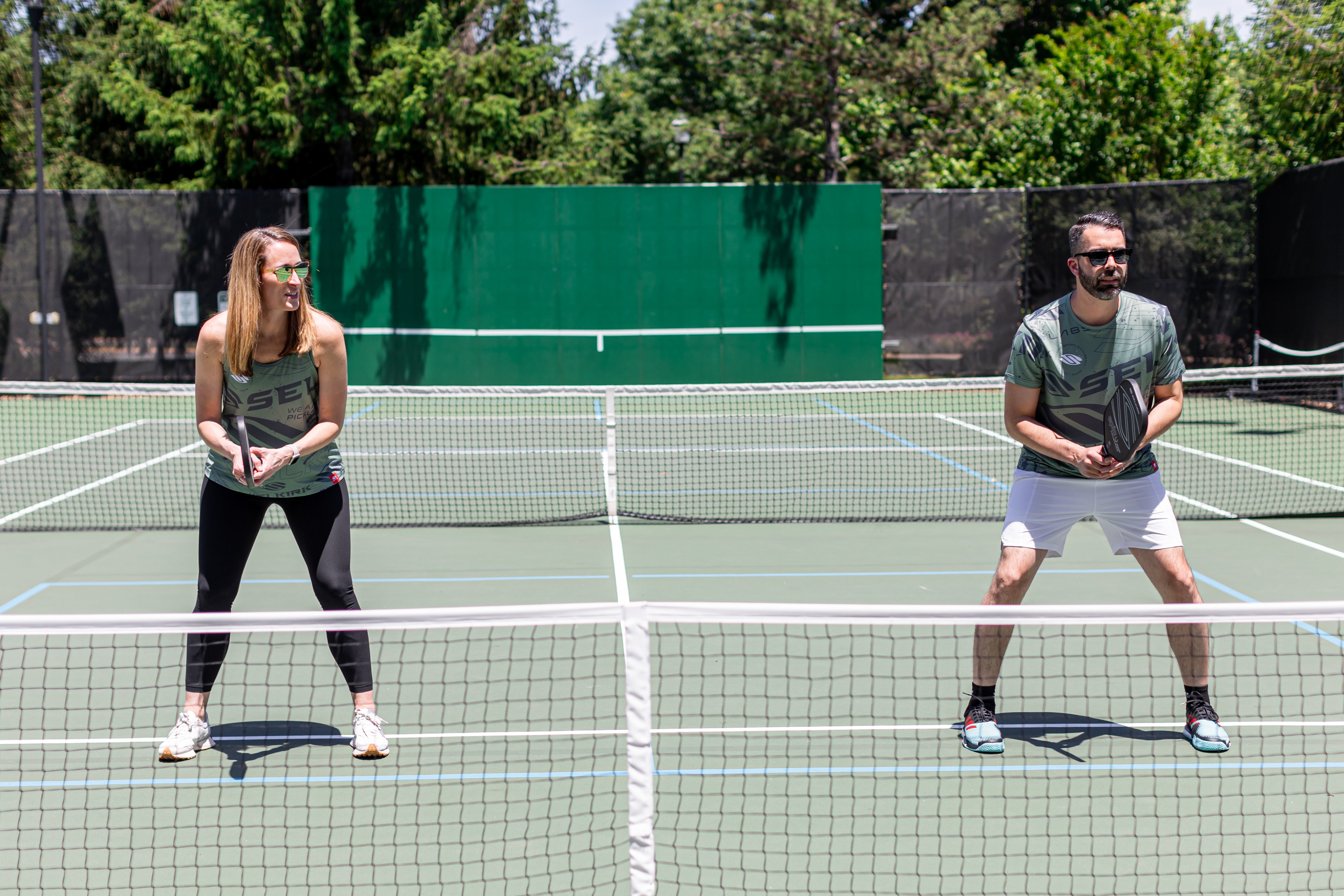 A woman and man stand at the kitchen line of an outdoor pickleball court, ready to receive a ball. 