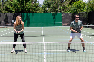 A woman and man stand at the kitchen line of an outdoor pickleball court, ready to receive a ball. 