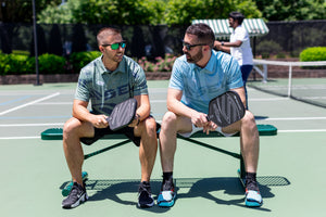 Two men sit on a bench near a pickleball court talking. 