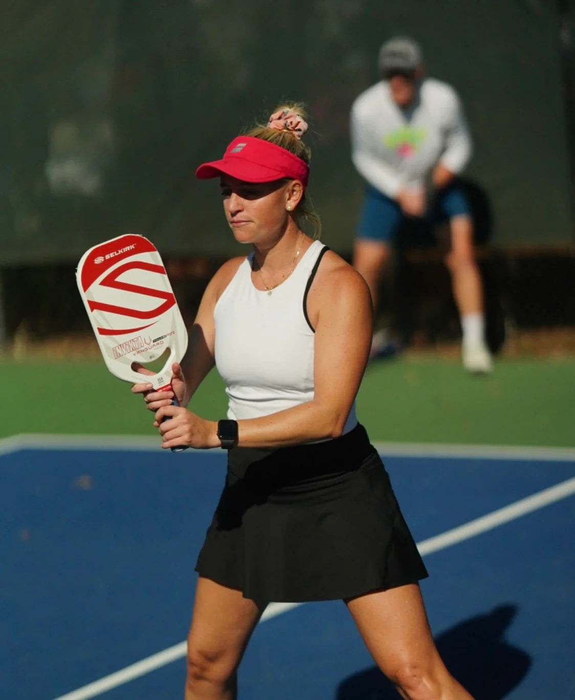 A female pickleball player in a white tank top and black skirt prepares to serve with a red paddle.