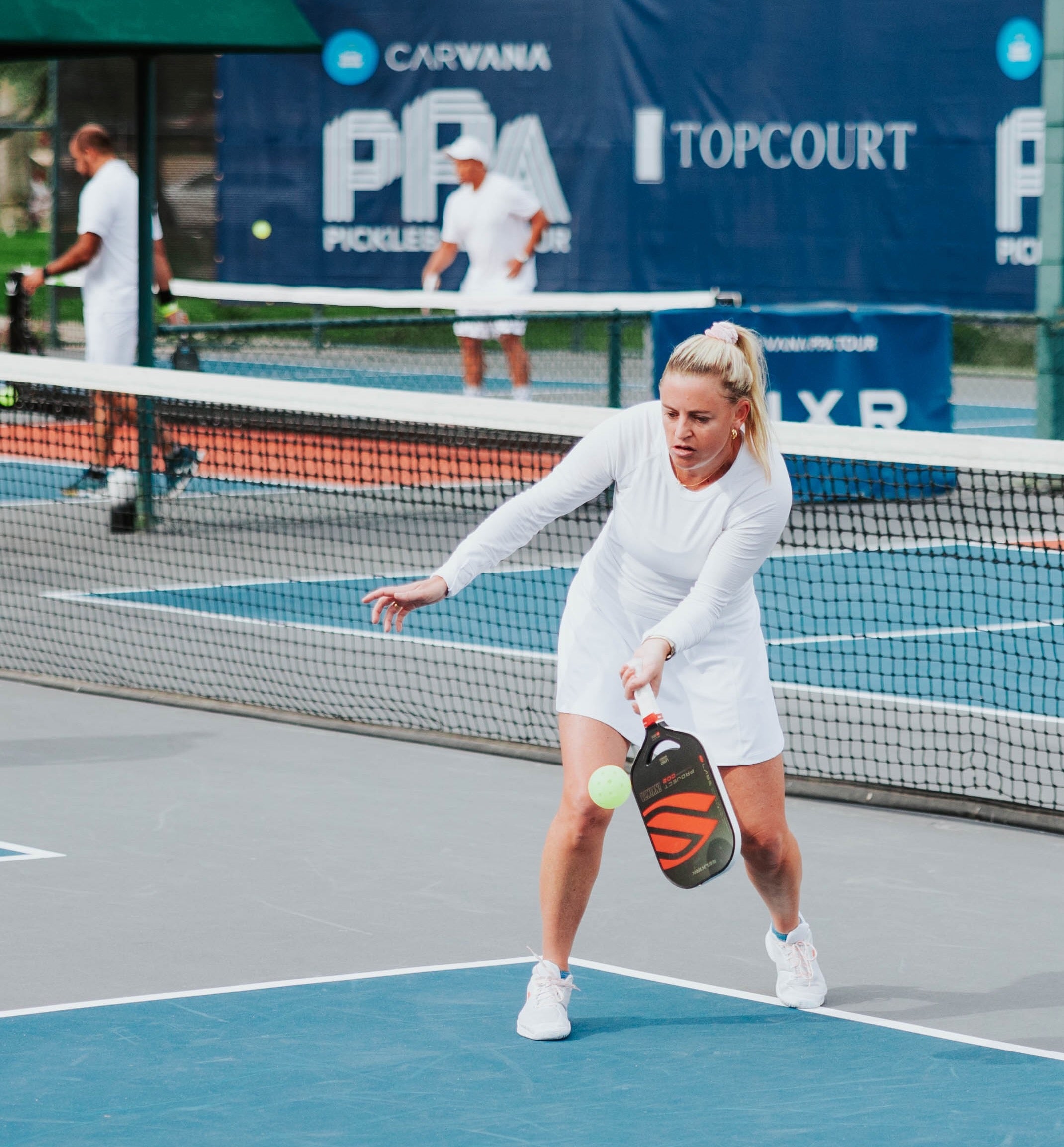 A female player in a white outfit executing a third shot drop in pickleball on a court.