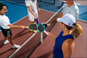Four players tap Selkirk paddles over a net after finishing a pickleball game. 