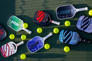 Several Selkirk paddles are lined up with pickleballs on an outdoor court. 