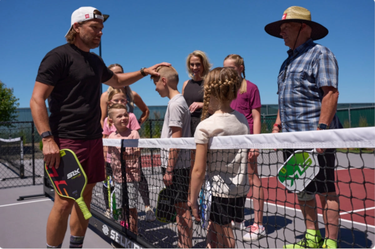 A group of children and adults interact near a net on an outdoor pickleball court, with paddles in hand.