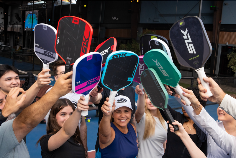 A group of pickleball players hold their SLK paddles up in the air. 