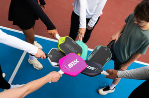 A group of pickleball players tap a variety of pickleball paddles on an outdoor court. 