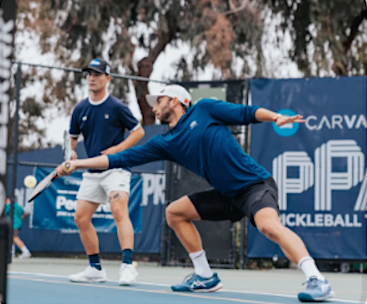 A man lunges to hit a pickleball while another player watches on a court with a pickleball banner in the background.