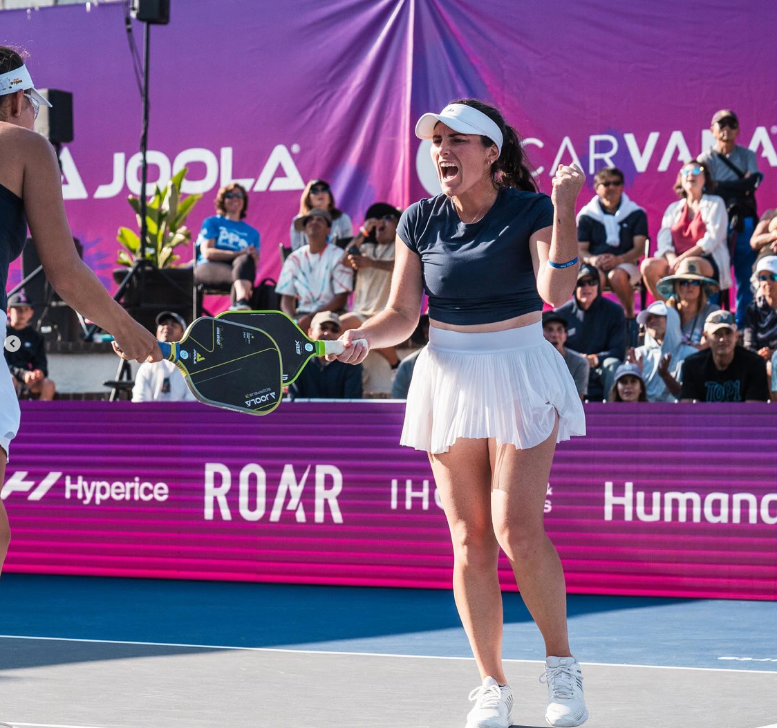 A female player celebrating with a partner during a pickleball match, with a cheering crowd in the background.