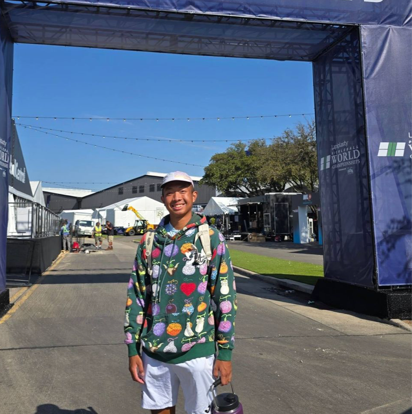 A young athlete wearing a colorful hoodie stands in front of a pickleball tournament entrance.