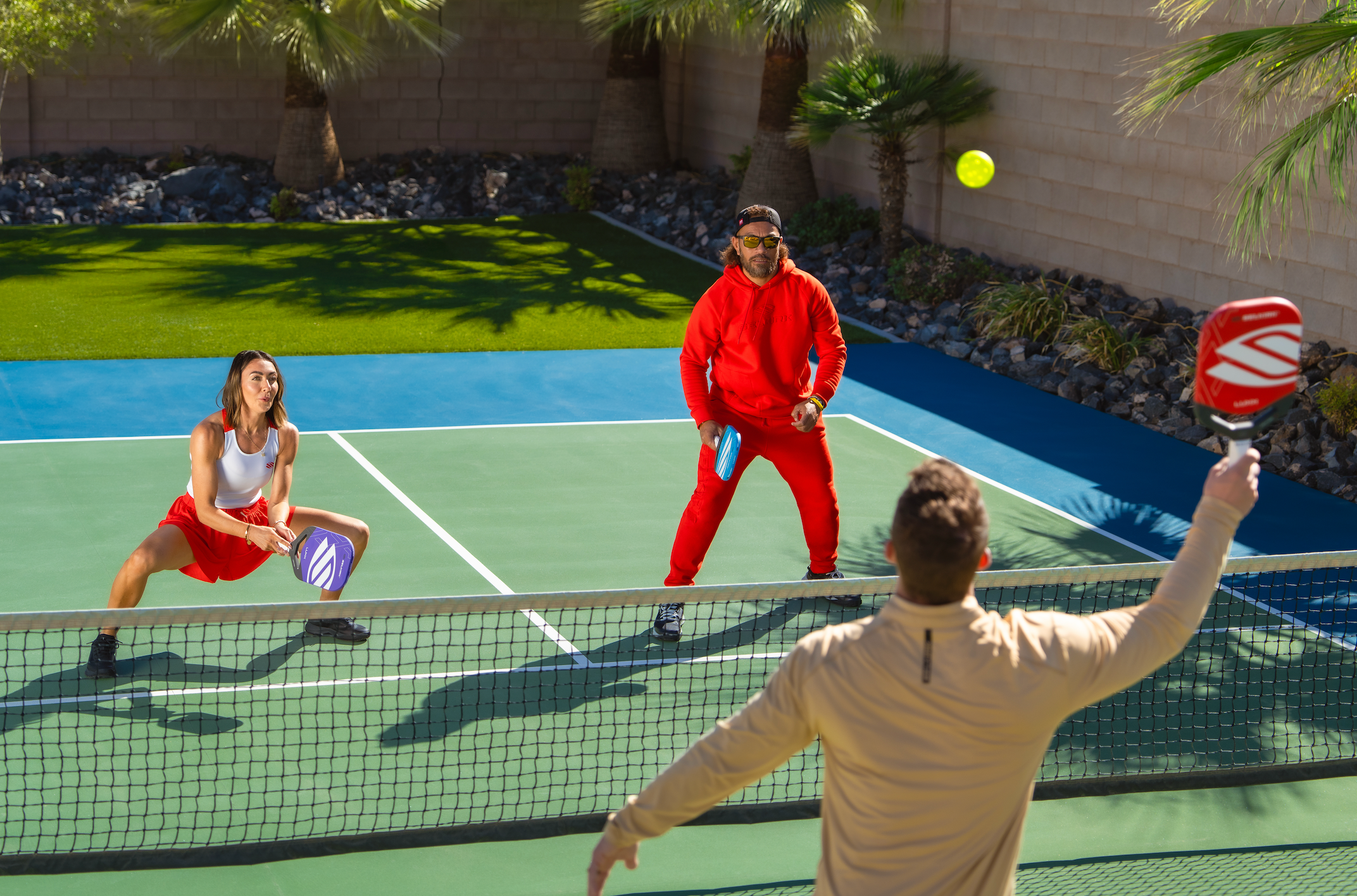Three people play pickleball on an outdoor court. A woman crouches low to receive an overhead shot a man is about to hit. 