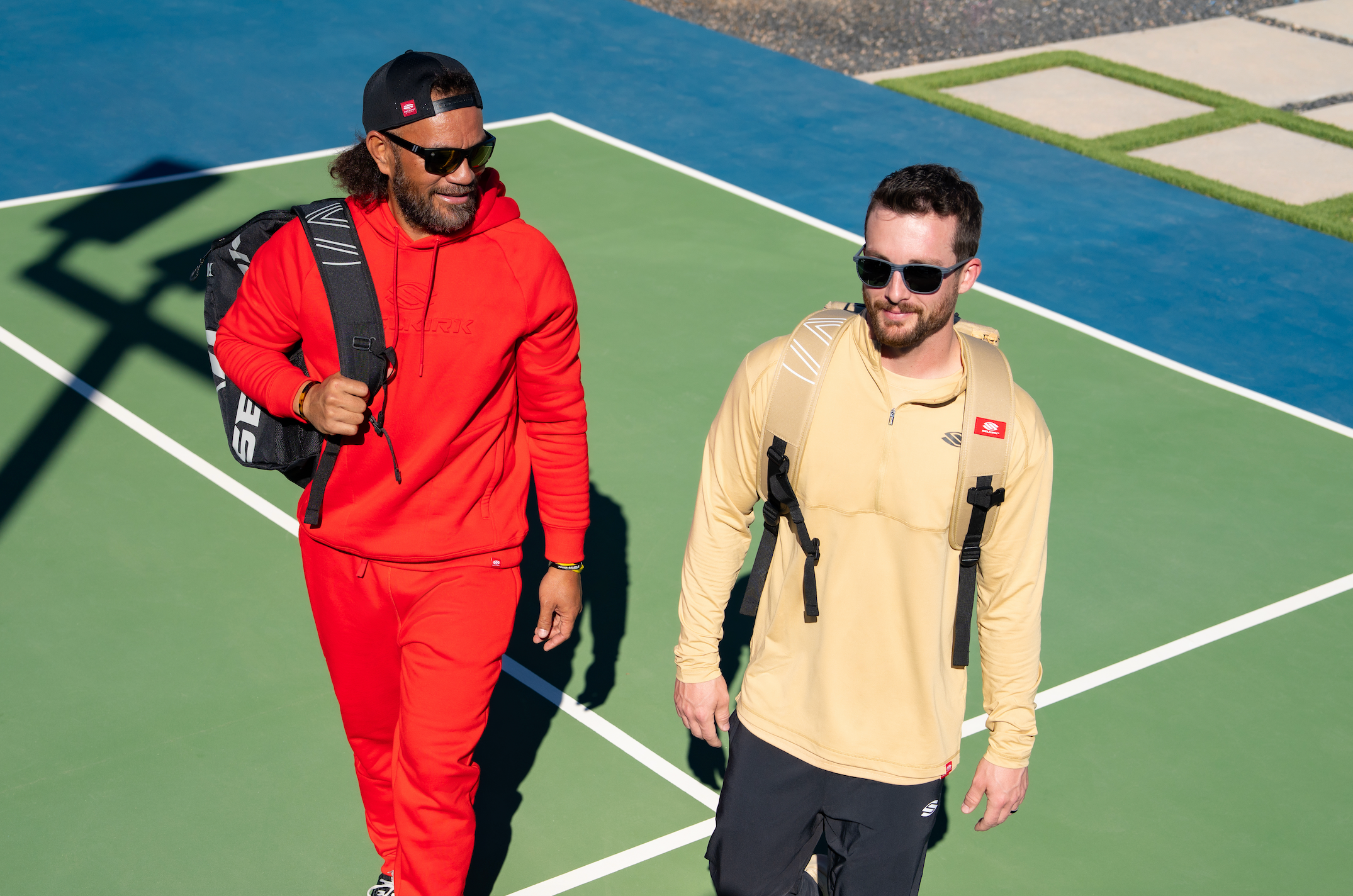 Two men walk onto an outdoor pickleball court with their Selkirk pickleball bags. 