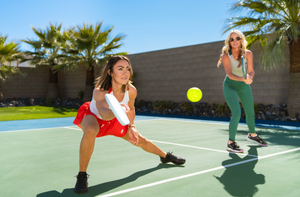 A woman dressed in red and white lunges for a pickleball shot while her partner watches. 