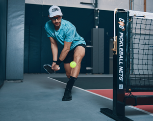 A man attempts to hit an around-the-post shot on an indoor pickleball court. 