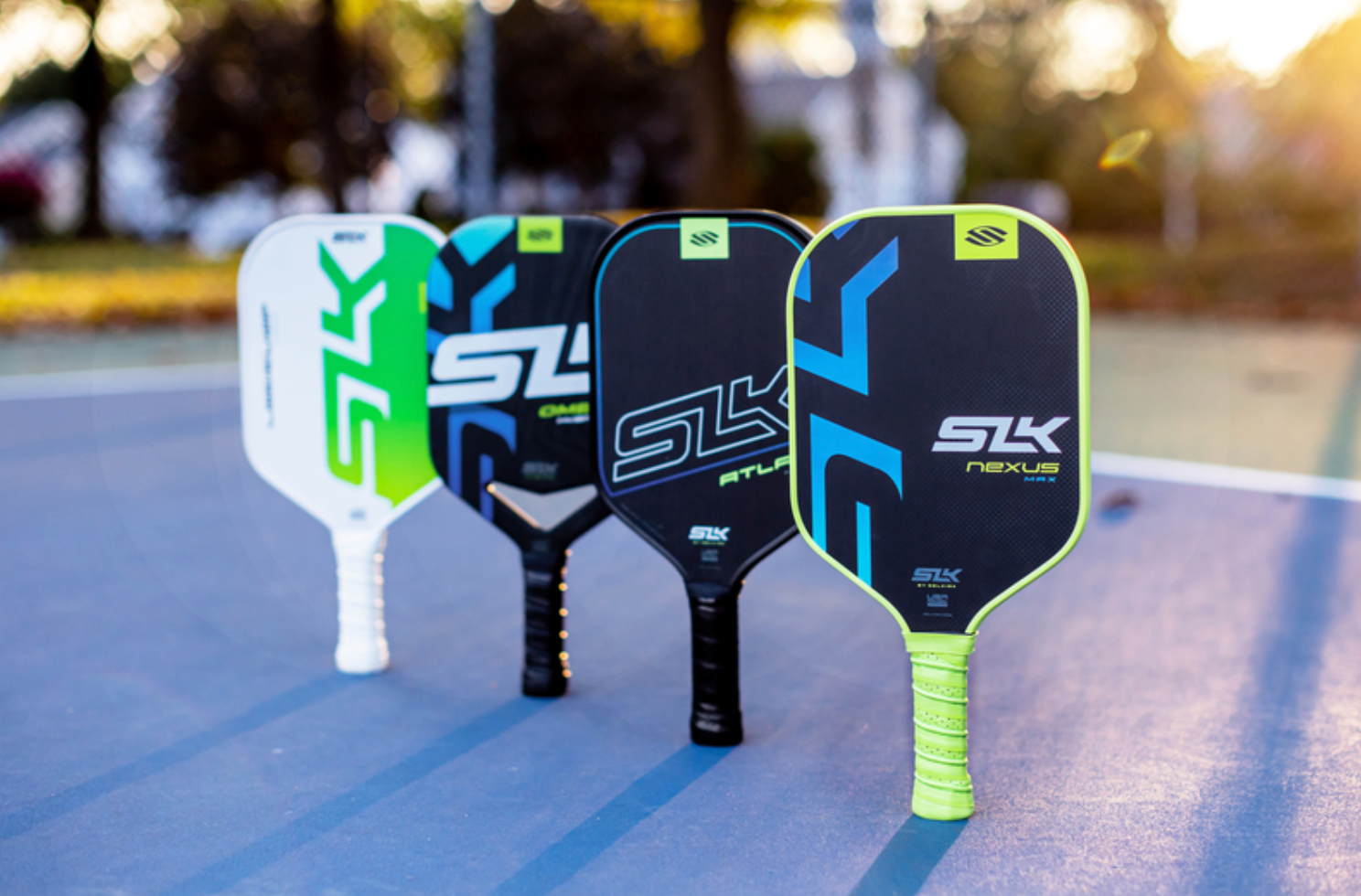 Four SLK paddles stand upright in a diagonal line on an outdoor pickleball court at sunset. 