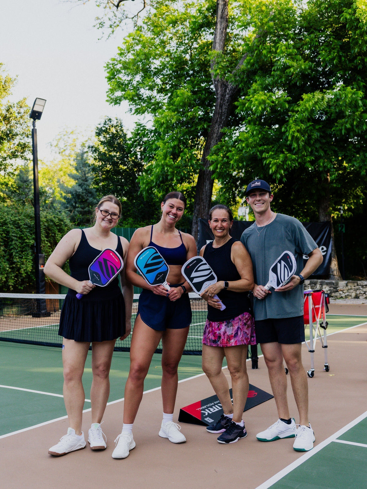 Four people smile as they hold their new Selkirk OMNI pickleball paddles on an outdoor court. 