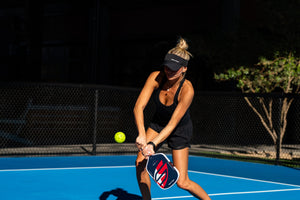 A woman wearing a visor hits a two-hand backhand third shot drop. 