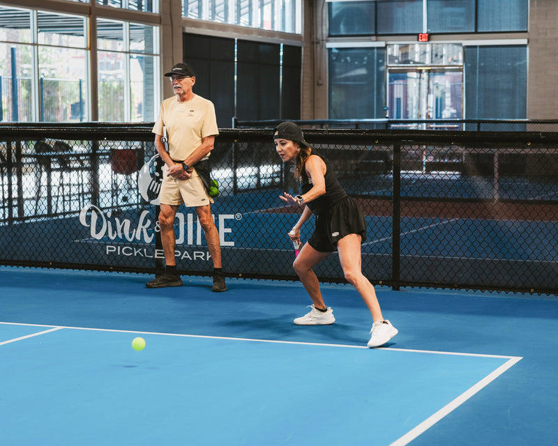 A woman, playing the right side of the court, steps in front of her partner to take the third shot with her forehand on an indoor pickleball court. 