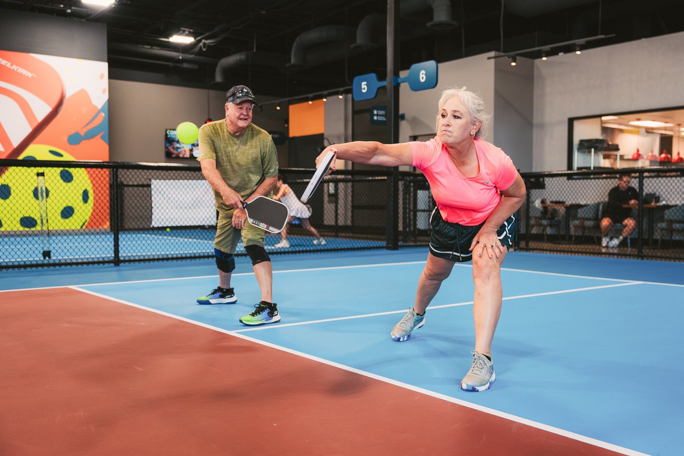 A woman performs a volley dink while lunging on an indoor court. Her partner looks on. 