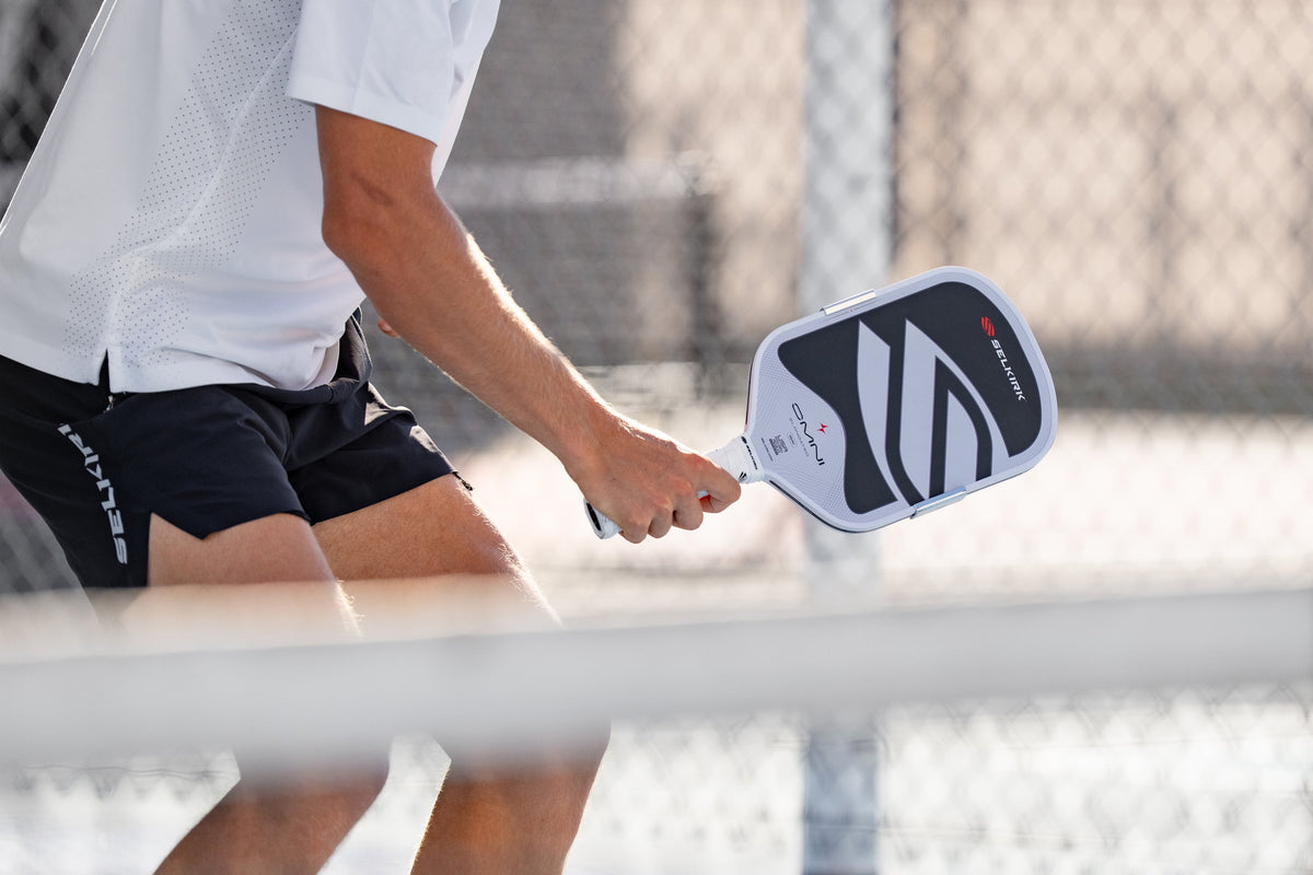 A close-up shows a man's arm as he holds a Selkirk OMNI all-court pickleball paddle. 