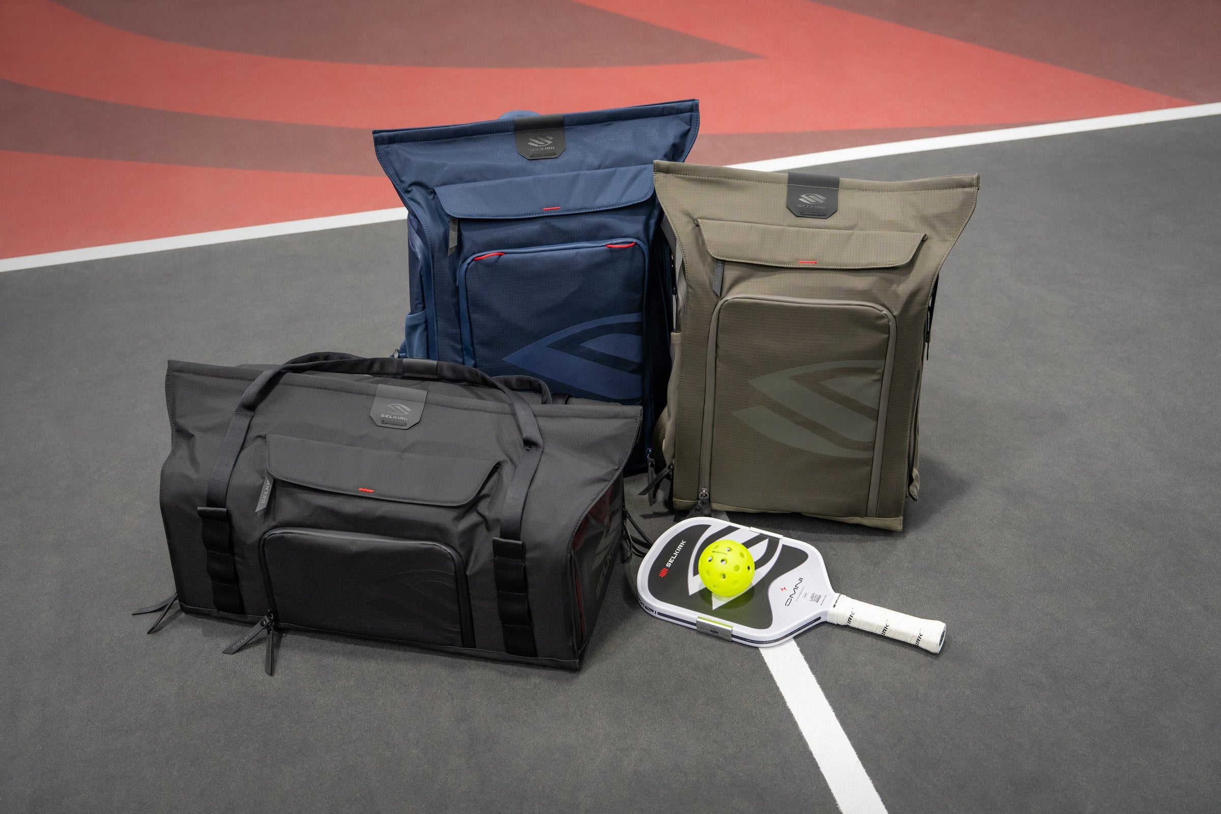 Three different sized pickleball bags sit next to each other on a pickleball court. 