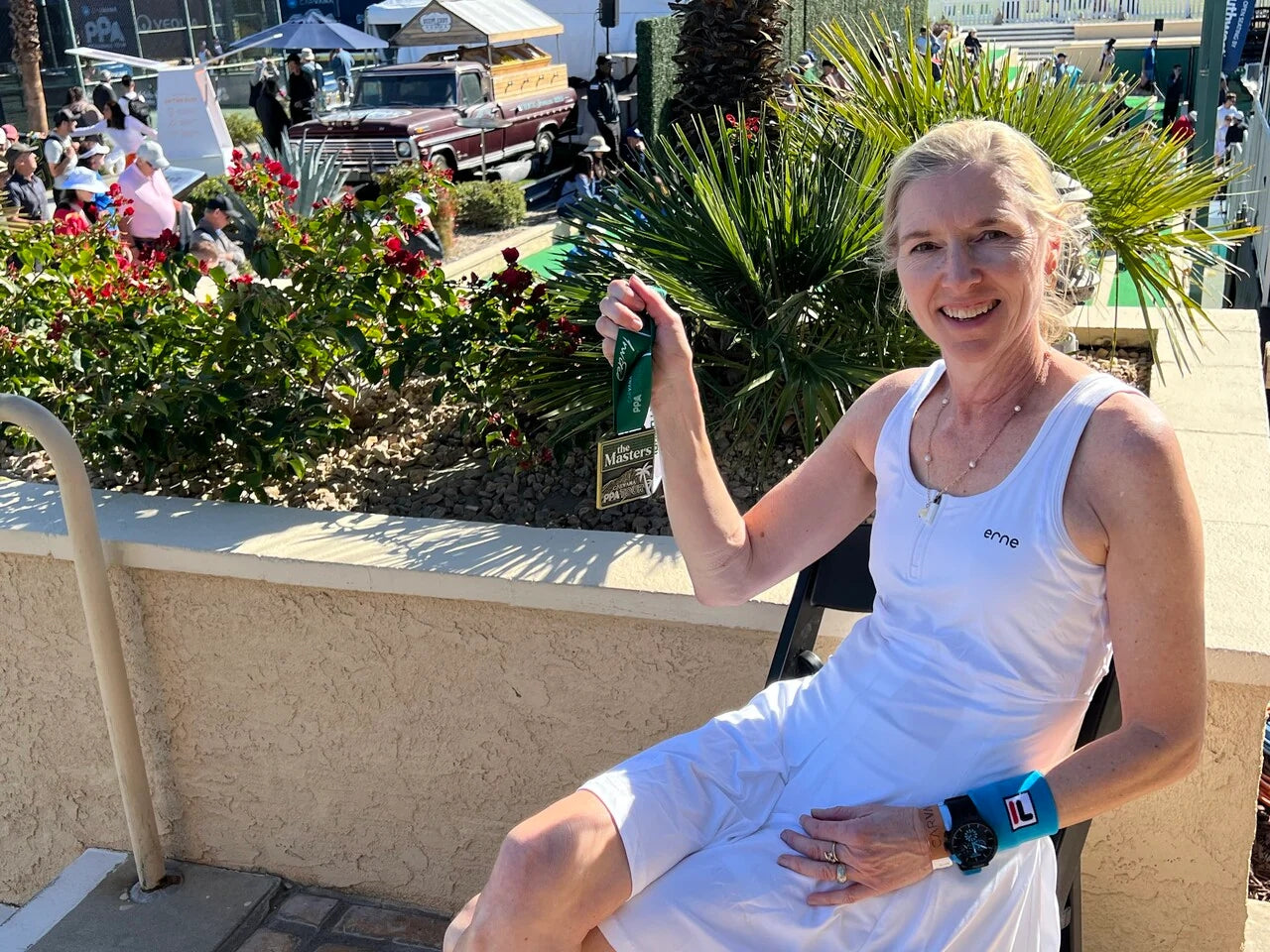 A woman in a white dress holding a pickleball medal while sitting outdoors.