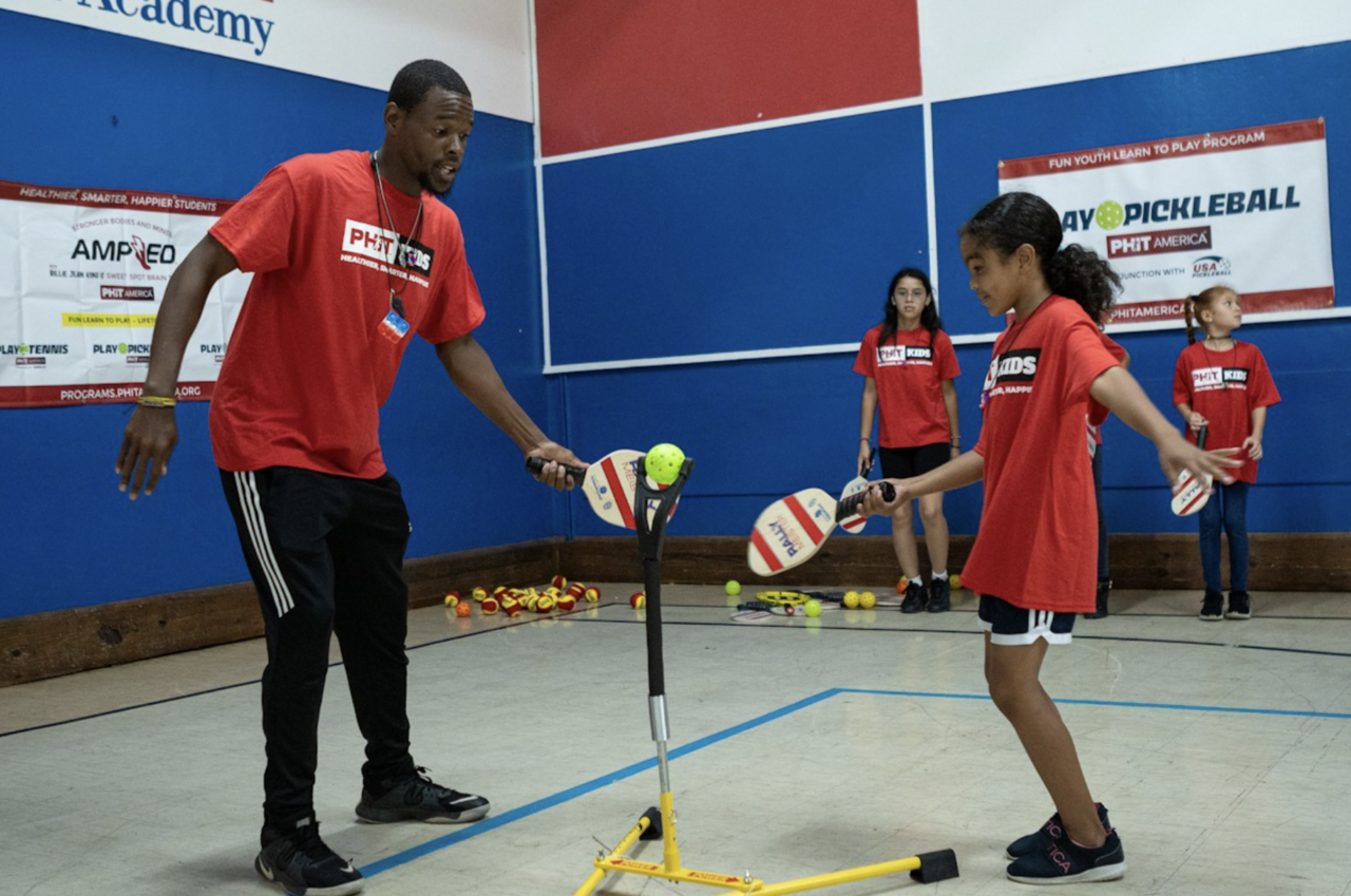 A young female student hits a pickleball off a tee while a male instructor looks on during a PHIT America pickleball course in a school gymnasium.