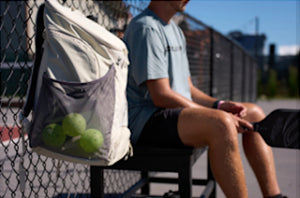 A white Selkirk pickleball backpack clipped to a fence with green pickleballs visible in the mesh pocket.