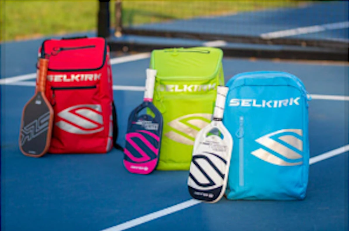 Three Selkirk pickleball bags in red, green, and blue with paddles on a pickleball court.