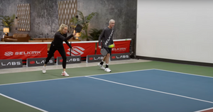 A woman serves a pickleball while she and her male partner begin to travel forward on an indoor pickleball court. 