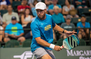 A man in a blue shirt and white cap playing pickleball, holding a paddle.