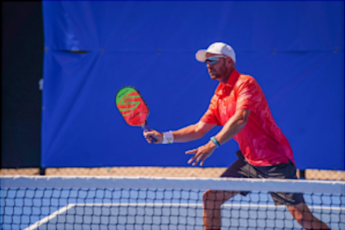 A player in a red shirt preparing to hit a pickleball with a paddle at a tournament.