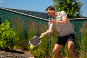 A young man performs a drop pickleball serve on an outdoor pickleball court. 