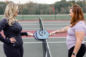 Two women smile as they tap Selkirk pickleball paddles after a match. 