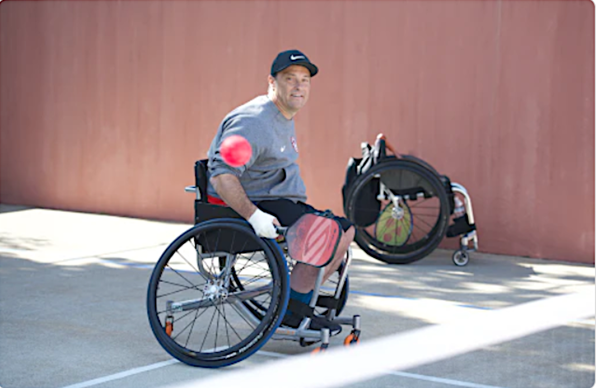 A man in a wheelchair smiles while preparing to hit a pickleball outdoors.