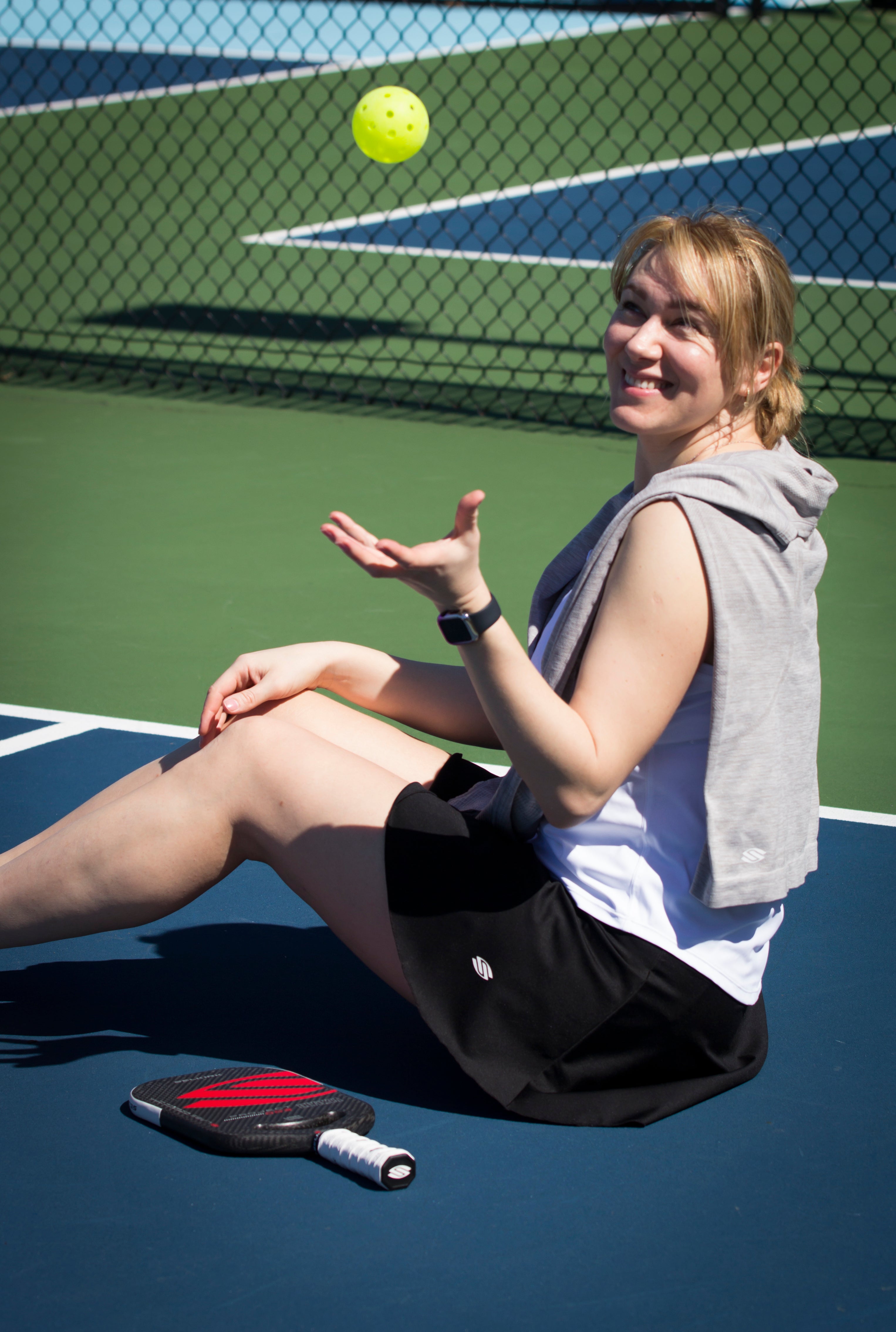 A woman sitting on a pickleball court, tossing a pickleball, wearing a gray hoodie and black skirt.